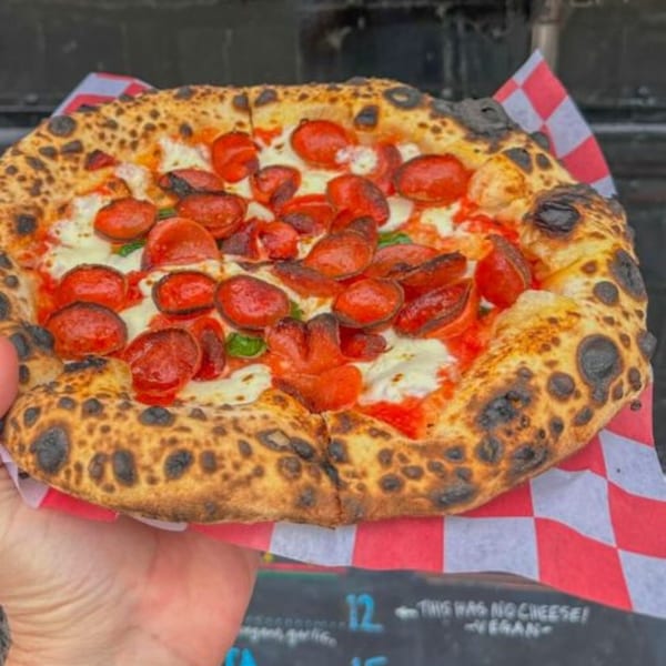 A hand holds a BareBakedPizza pepperoni pizza with a charred crust over red and white checkered paper.