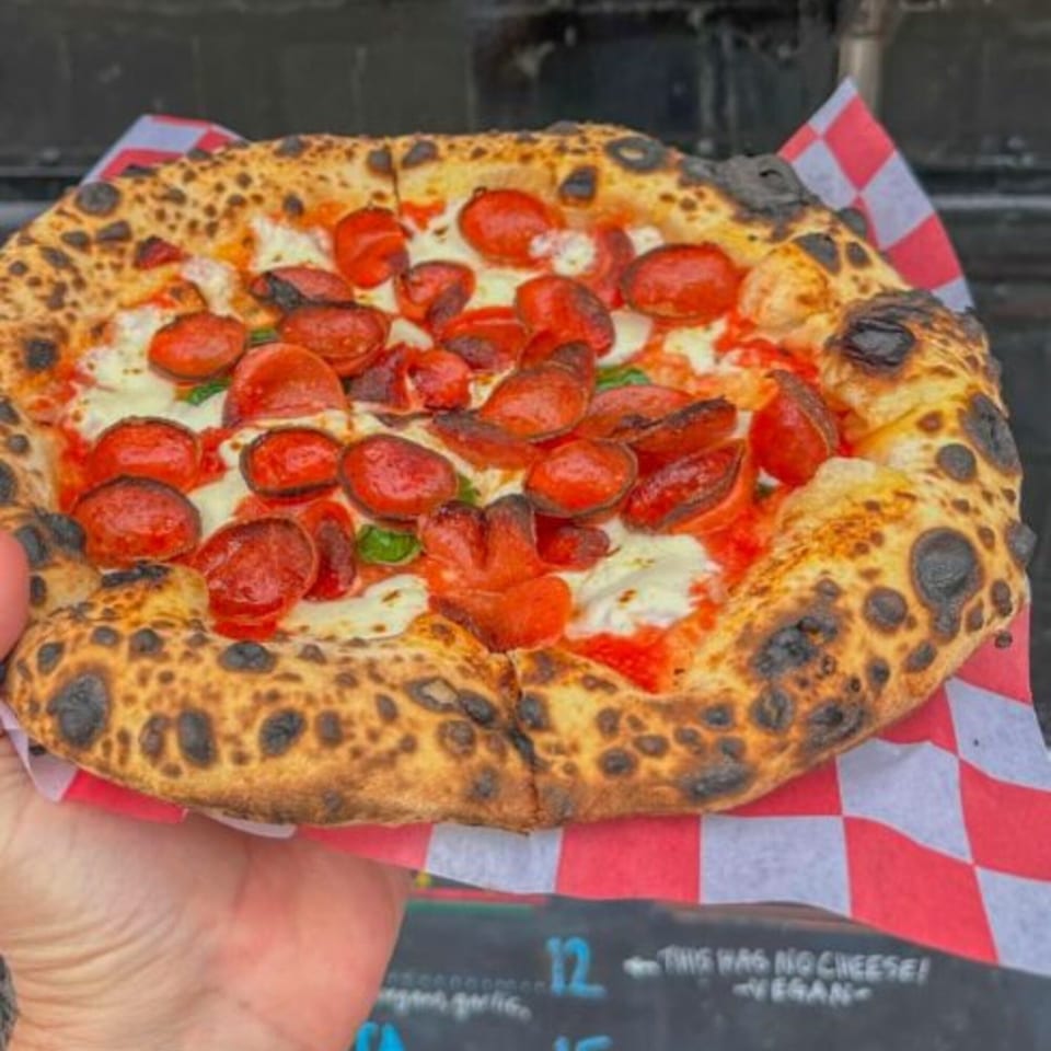 A hand holds a BareBakedPizza pepperoni pizza with a charred crust over red and white checkered paper.